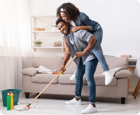 A man gives a piggyback ride to a smiling woman while he holds a mop in a clean room.